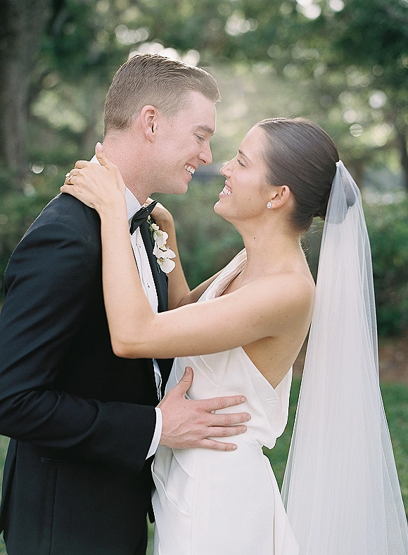 Couple portrait of bride and groom embrace with a forehead touch, her veil and strapless dress against sunlit greenery bokeh
