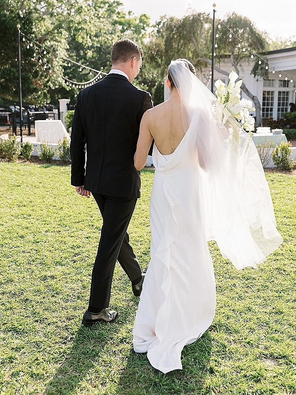 Wedding couple walking hand in hand, bride and groom from behind, her long veil and calla lily bouquet on a garden lawn under string lights