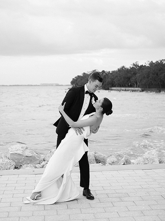 Wedding couple portrait in a dip kiss pose, bride in strapless dress and heels with groom in black tux by rocky waterfront walkway