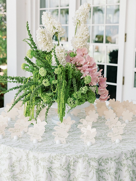 Escort card display with wedding escort cards in butterfly designs around a lush white and pink orchid centerpiece on a porch table by French doors