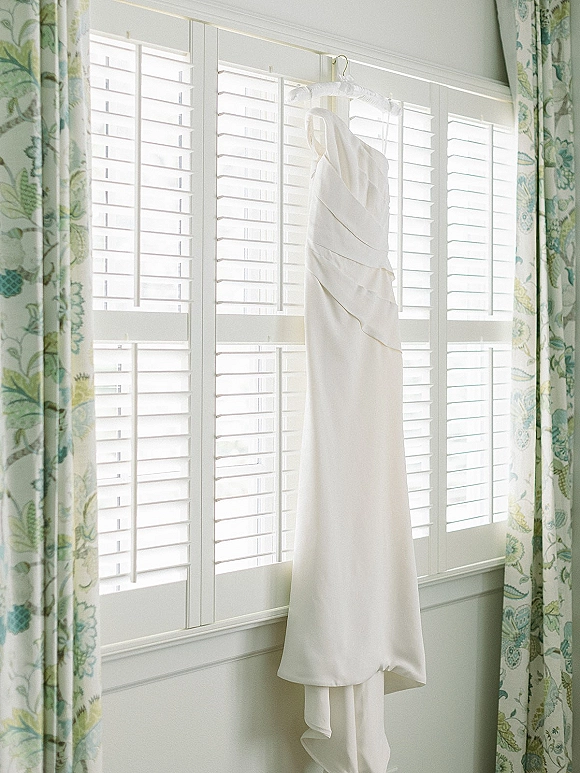 Wedding dress on hanger glowing in window light, framed by floral curtains and white shutters against classic wall paneling