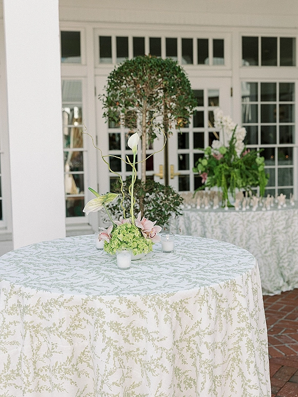 Cocktail table decor with botanical tablecloth, orchid and hydrangea centerpiece, and glass votive candles on a brick patio by white French doors