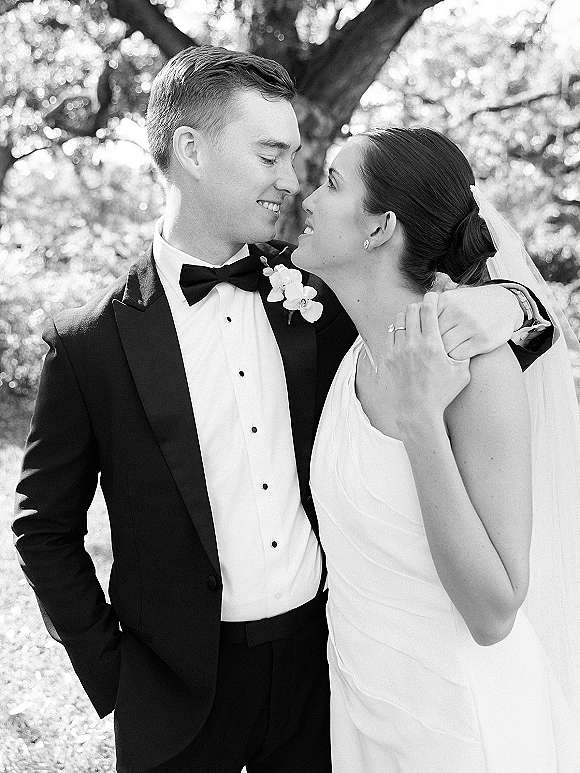 Couple portrait in a black and white wedding portrait, bride in veil and strapless dress embracing groom in tuxedo amid sunlit garden foliage