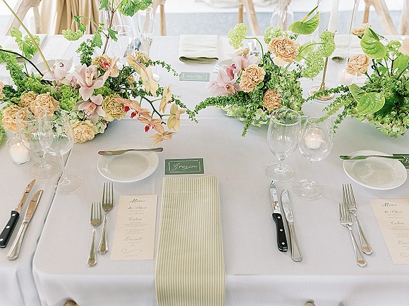 Reception tablescape with a wedding floral centerpiece of orchids, peach carnations, and hydrangea, set with brass candles, menus, and striped napkins