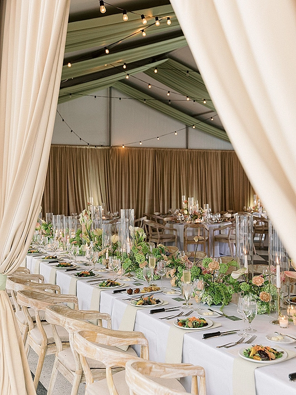 Reception tablescape with long head table setup, white linens, greenery garland and pastel flowers, taper candles under string lights in a tent