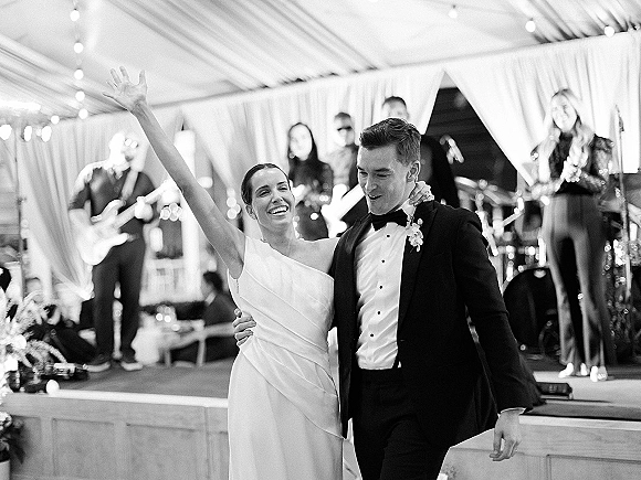 First dance moment as bride in a wedding dress raises her arm with groom in tuxedo under string lights in a wedding tent, band on stage behind