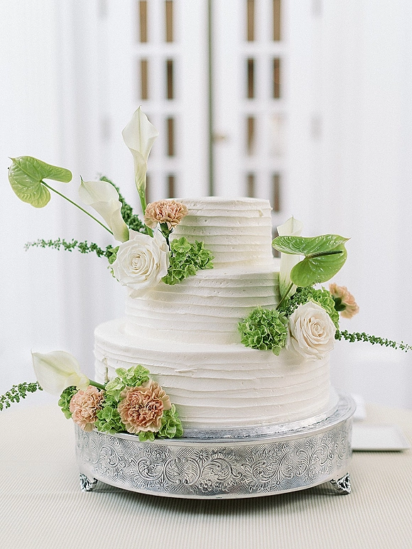 Wedding cake with white buttercream in a two tier wedding cake design, topped with roses and calla lilies on an engraved silver pedestal backdrop