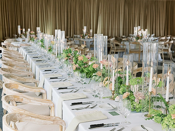 Reception tablescape with white linens, floral garland and taper candles in glass hurricane vases, set in a draped-curtain reception room