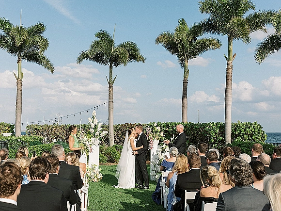 Ceremony kiss as bride and groom embrace under white arch columns with floral pillars, string lights, and ocean palms beyond chairs