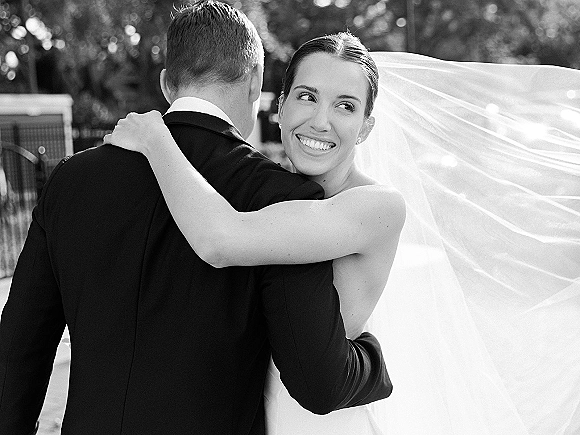 Wedding couple portrait of a bride hugging groom over her shoulder, smiling as her veil blows, with trees and a fence behind them