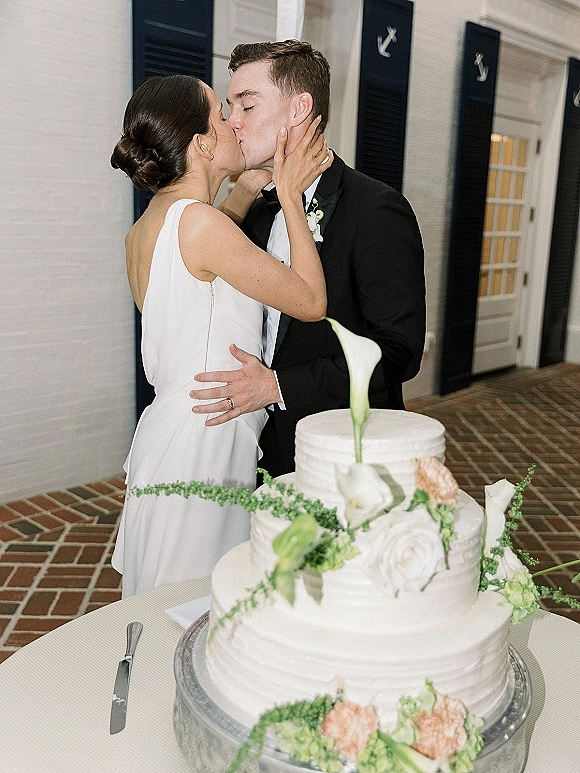 Wedding kiss beside a white buttercream cake with calla lilies and greenery garland, on a brick floor against a white brick wall