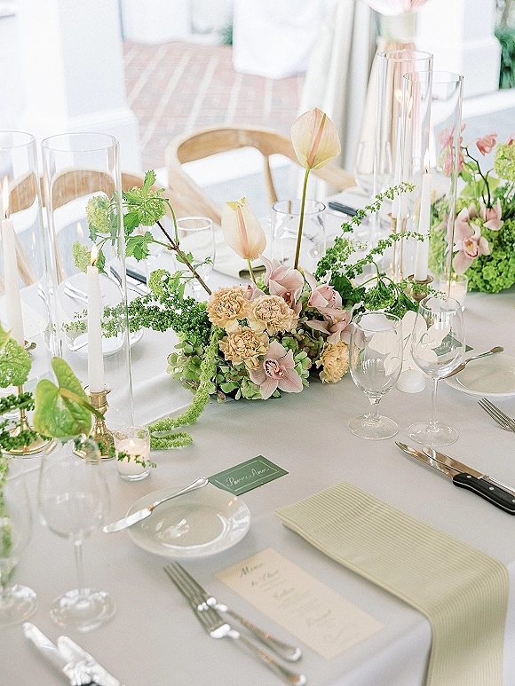 Reception tablescape with wedding table centerpiece of pastel florals, taper candles, bud vases, and place cards under white tent drapery on brick floor