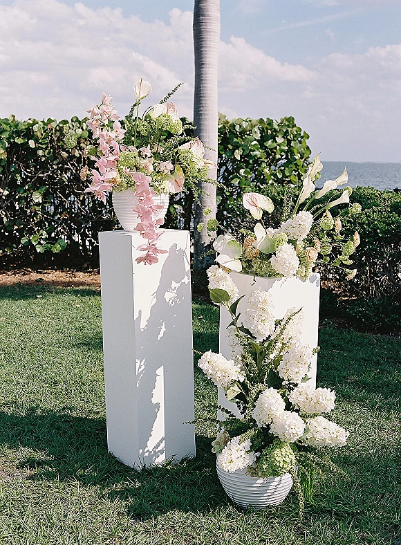 Wedding floral arrangement on white pedestal plinths with calla lilies and hydrangeas, set on a coastal lawn with ocean backdrop