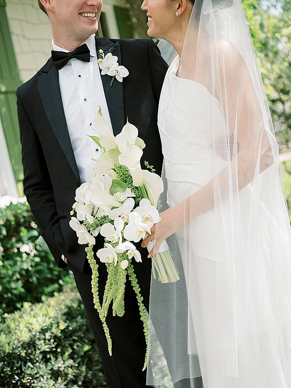 Couple portrait of bride and groom smiling, her veil and orchid bouquet beside his black tuxedo, outside a sunlit house with green trim