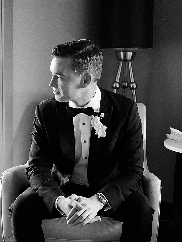Groom portrait of a black and white groom portrait seated in an armchair, wearing tuxedo with bow tie and boutonniere near a floor lamp