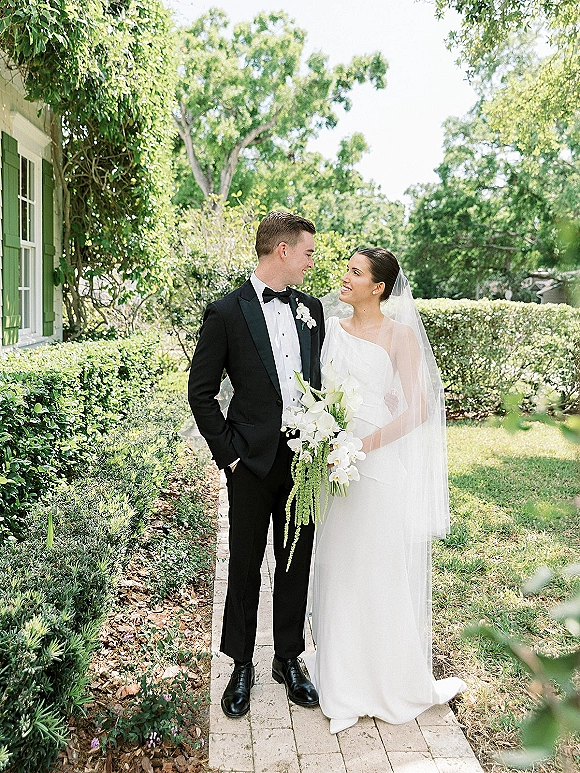 Couple portrait of bride and groom looking at each other, her long veil and white calla lily bouquet on a brick garden path