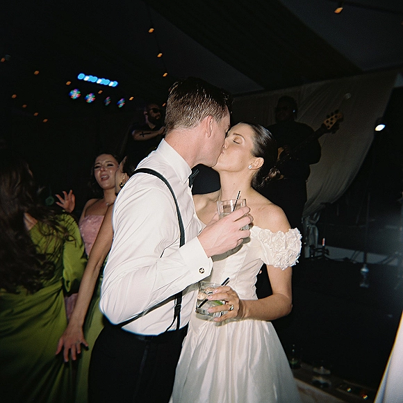 Wedding kiss as bride and groom kiss on a lively dance floor, each holding cocktails, with guests and band under stage lighting