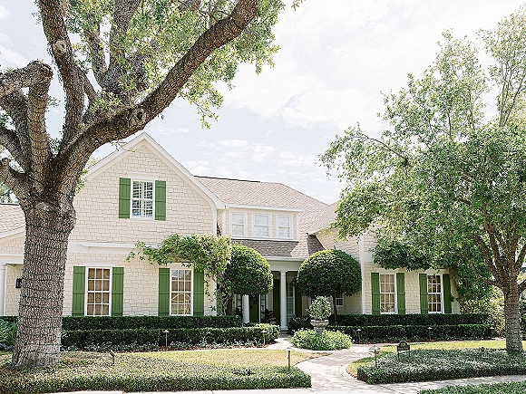 Wedding venue exterior with a white house and green shutters, framed by manicured hedges and topiary trees along a lit walkway to the porch