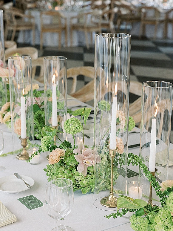 Reception tablescape with wedding table centerpiece of hydrangeas and orchids, taper candles in hurricanes, gold candlesticks on white linen