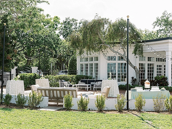 Outdoor reception lounge with wedding lounge seating under string lights, featuring sofas, cocktail tables, and a beverage station on a patio lawn