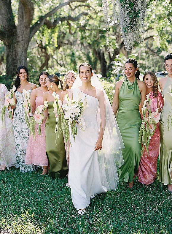 Bride with bridesmaids in a bridal party portrait walking on a lawn under oak trees, wearing veil and carrying orchid calla lily bouquets