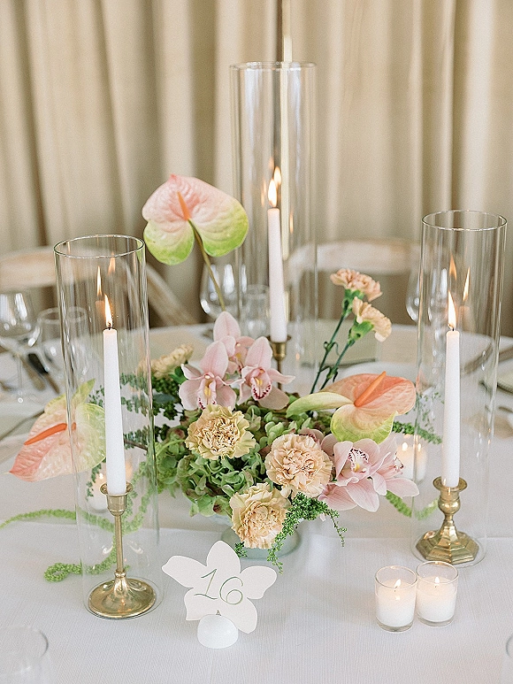 Wedding centerpiece with a wedding floral centerpiece of anthurium and orchids, brass candlesticks and glass hurricanes on a white tablecloth table