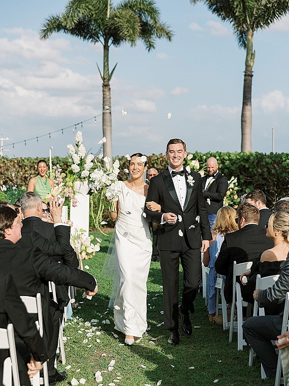 Wedding recessional as bride and groom walk the aisle amid a rose petal toss, bouquet and veil flowing under palm trees and string lights