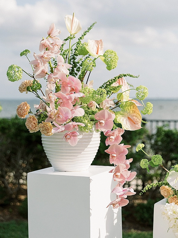 Wedding floral arrangement with pink orchids and anthurium in a white ceramic vase on a plinth, set against ocean horizon and hedges