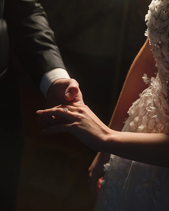 Wedding ring close-up of bride and groom holding hands, engagement ring sparkling beside lace cuff and suit sleeve against dark background
