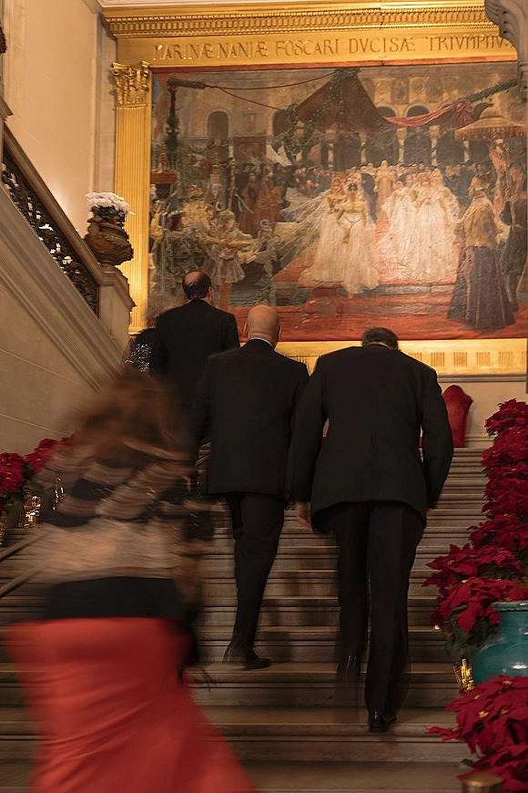 Wedding guest attire with black tie wedding guests in tuxedos and a red dress ascending a grand staircase beneath a mural and urns