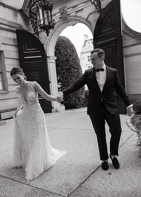 Couple portrait of bride and groom holding hands, walking past a stone archway entrance with a lace wedding dress accent