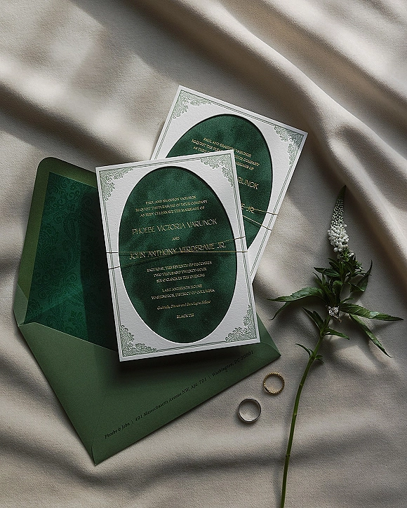 Wedding invitation suite with emerald green envelopes and gold foil lettering, twine, floral sprig, and rings on a linen backdrop