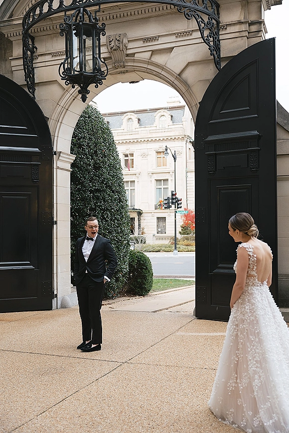 First look moment as the bride in an open-back lace gown approaches the groom in a tuxedo under a stone archway at the venue entrance