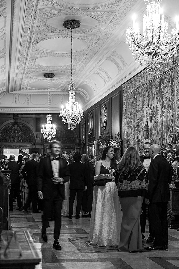 Wedding cocktail hour with black tie wedding guests mingling by a cocktail table under crystal chandeliers in an ornate ballroom