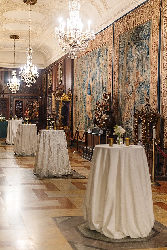 Cocktail hour setup with wedding cocktail tables draped in white linens, floral centerpieces, and votive candles in an ornate hallway under chandeliers