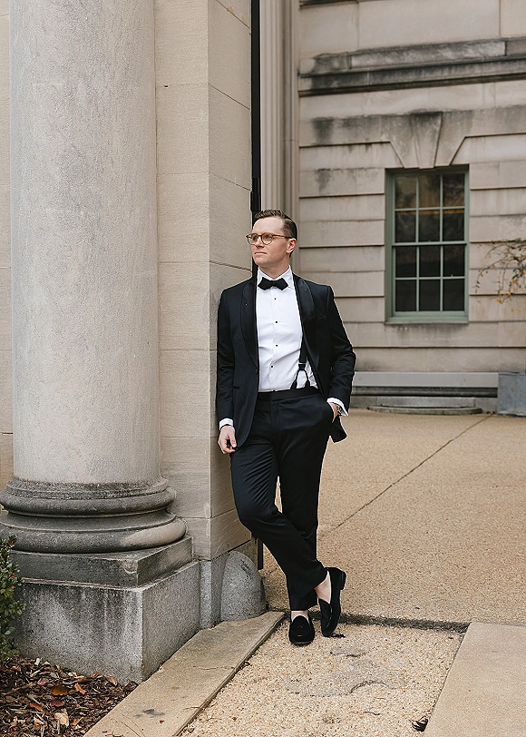 Groom portrait in a black tuxedo with bow tie, suspenders, and glasses, leaning by a stone column along a walkway outside a building facade