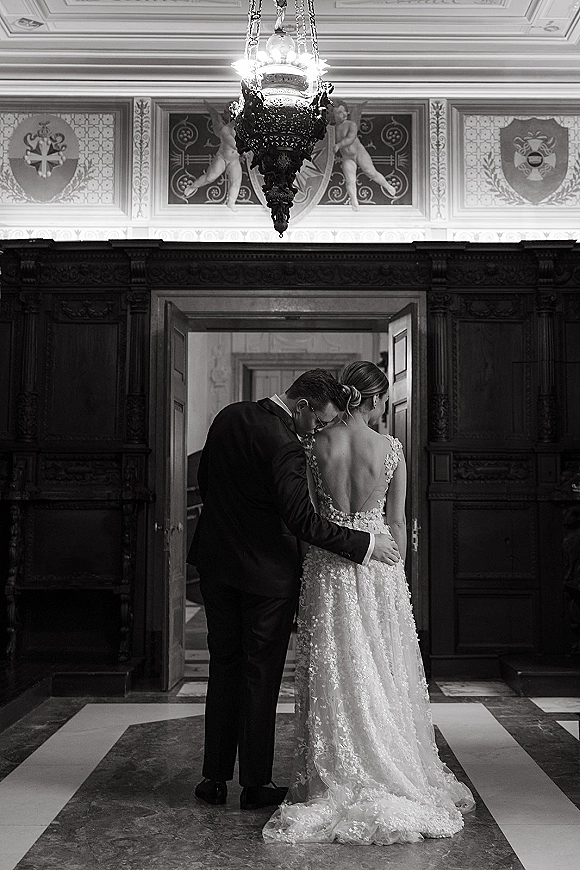 Couple portrait in a black and white wedding portrait, bride in a backless lace gown and groom in suit under a chandelier in a wood hallway