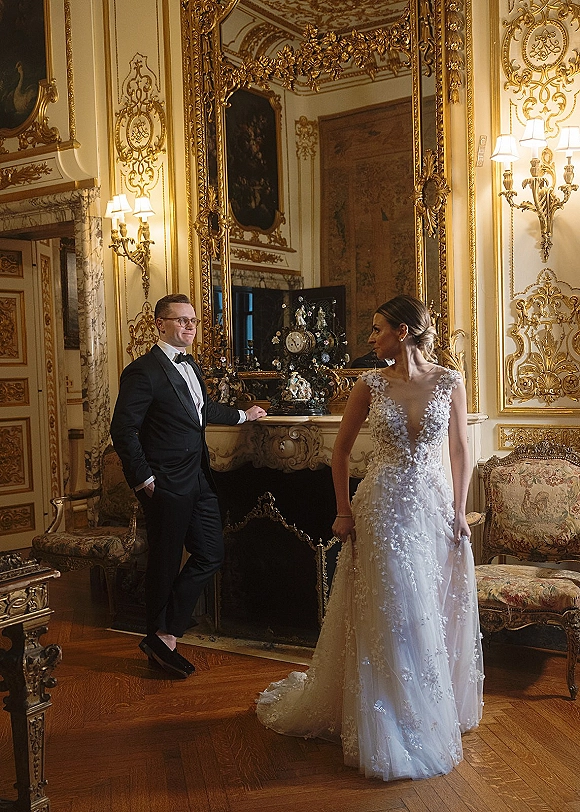 Couple portrait of bride in lace applique wedding dress and groom in tuxedo by a marble fireplace, reflected in an ornate gold mirror.