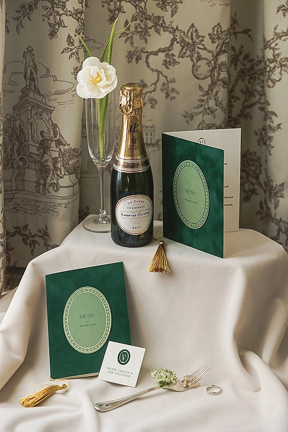 Wedding stationery flat lay with emerald green wedding invitations, menu and place card, champagne flute, ring, gold tassels on linen
