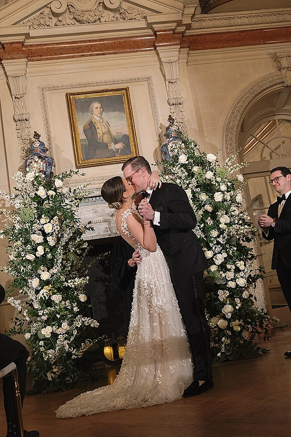 Wedding kiss portrait of bride and groom kissing beneath a white rose arch with candles, set before an ornate fireplace mantel indoors