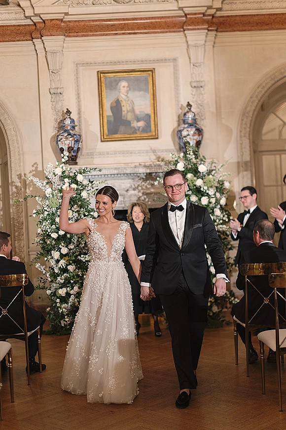 Wedding recessional as bride in lace gown waves while holding groom’s hand, walking past gold chairs beneath a white floral arch indoors