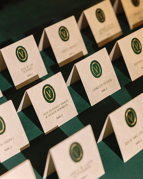 Wedding place cards in green ink and gold foil with a monogram crest on patterned cardstock, displayed on a dark green board