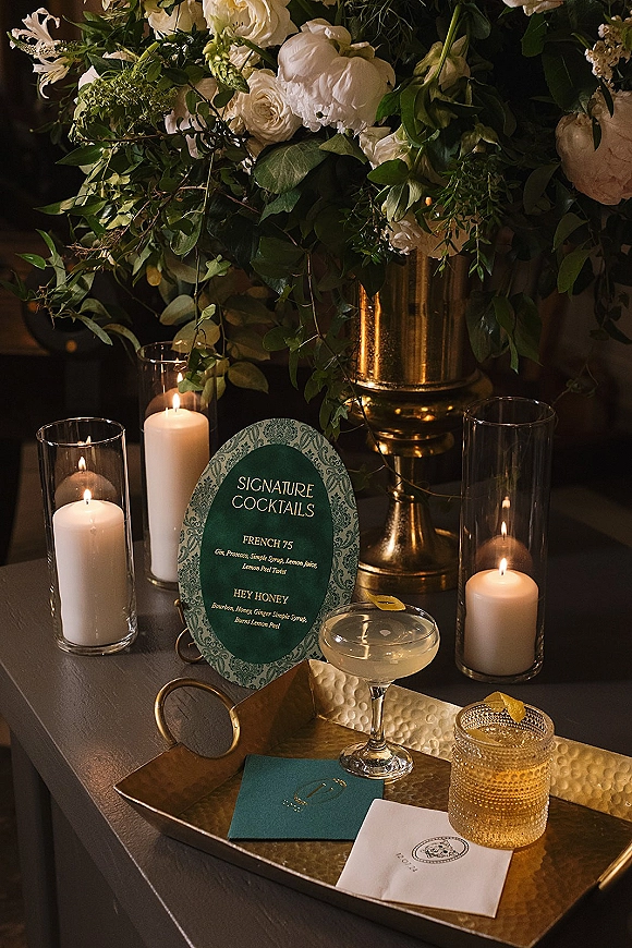 Wedding bar decor with a signature cocktail sign beside candlelit glass hurricanes, gold vase florals, and coupe and rocks glasses on the counter