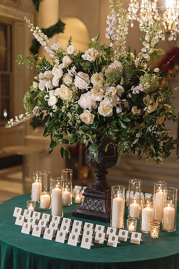 Escort card table with wedding escort cards display, white rose and greenery urn centerpiece, pillar and votive candles under a chandelier indoors