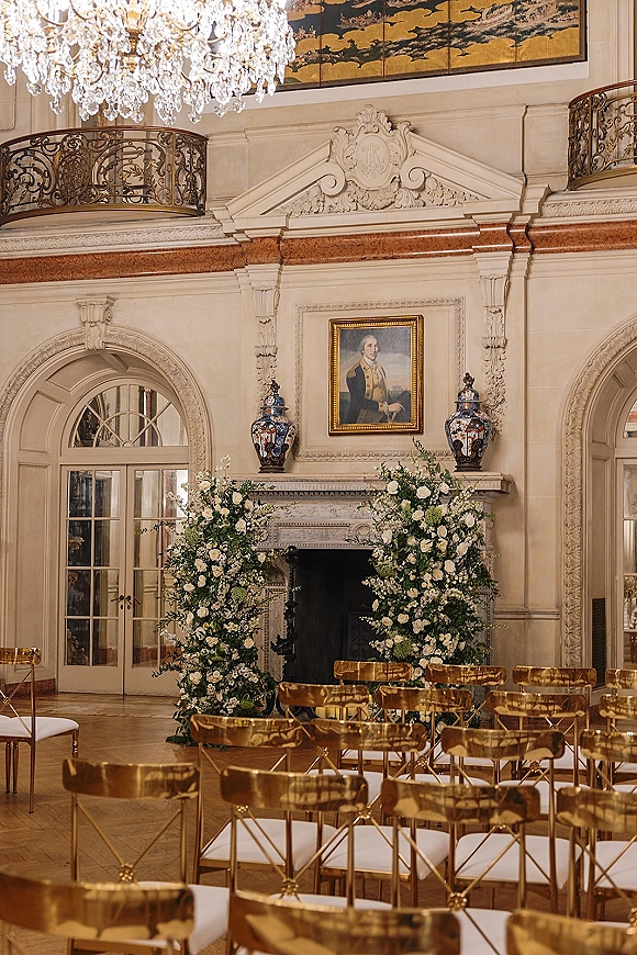 Indoor ceremony setup with white floral and greenery pillars framing a fireplace altar, gold chairs under a chandelier in a grand hall