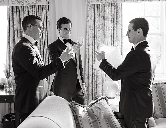 Groomsmen toast as men in tuxedos and bow ties clink champagne flutes in a bright sitting room by the window, laughing together