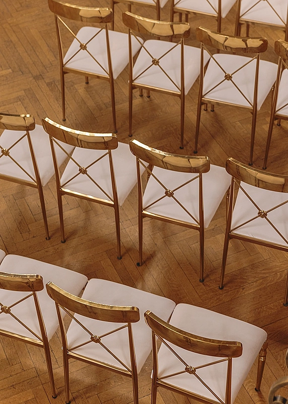 Ceremony seating with gold wedding chairs and white cushions arranged in neat rows on a wood parquet floor indoors