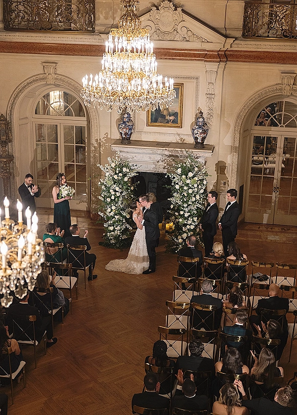 Ceremony kiss as the couple shares their first kiss at altar beneath a floral arch in a candlelit ballroom with chandelier and guests seated