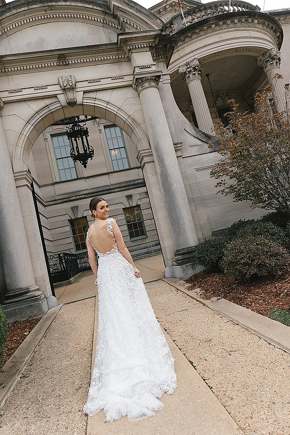 Bridal portrait of a bride in a backless wedding dress with lace straps and train, looking over her shoulder by a stone arched entryway