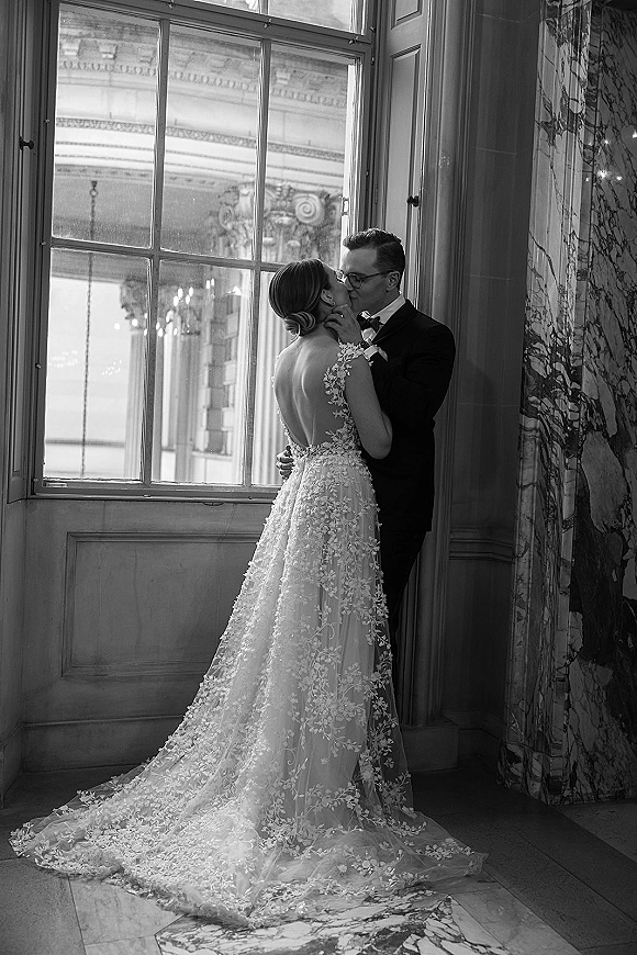 Wedding kiss portrait of bride and groom kiss by a large window, her lace gown with long train and floral appliqués beside his tuxedo and glasses